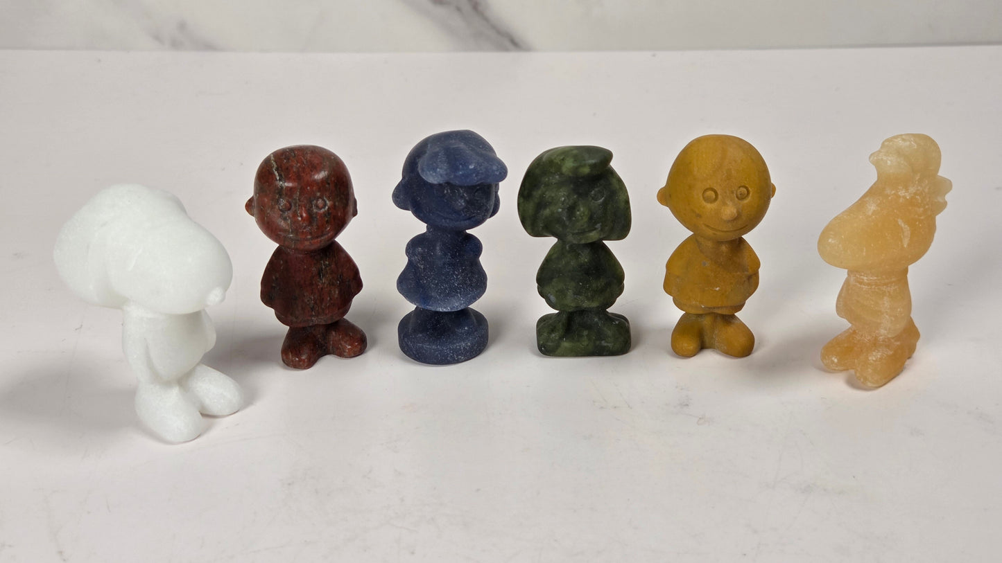 Six small carved figurines made from natural stone in various colors, arranged in a row on a white marble surface against a white marble background.