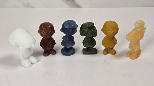 Six small carved figurines made from natural stone in various colors, arranged in a row on a white marble surface against a white marble background.