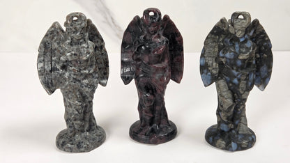 Three small angel figurines made from polished stone, displayed side by side on a white surface against a light gray marble-patterned background. The left figurine is made of gray and black marbled stone, the center figurine is made of dark reddish-brown and black marbled stone, and the right figurine is made of dark gray and blue marbled stone.