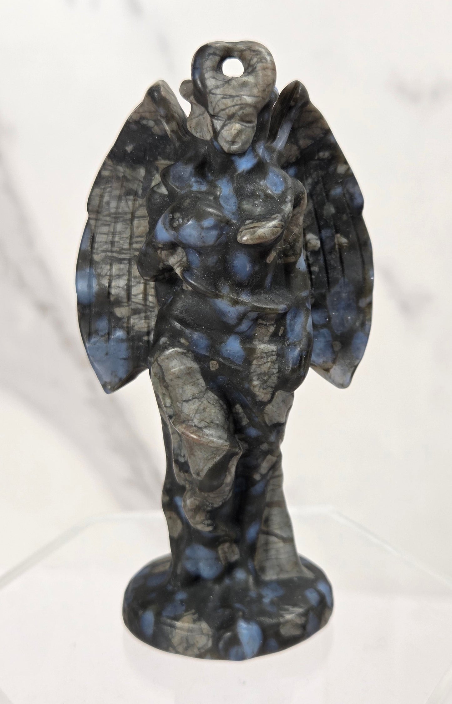 A small winged angel or cherub figurine made from mottled dark stone with blue and gray marbling, standing on a clear display base against a light marble-patterned background.