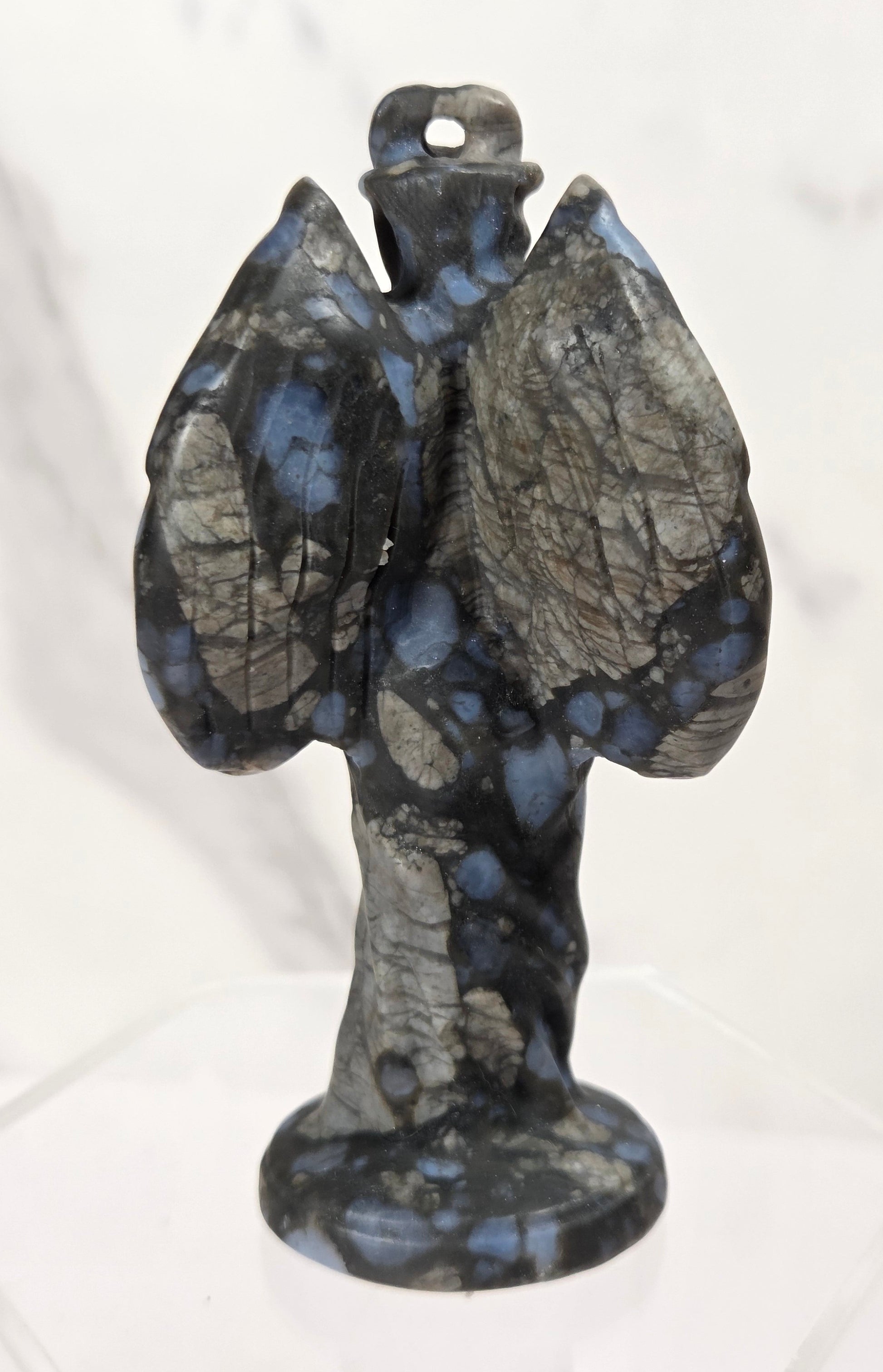 A small carved stone angel figurine with wings spread, standing on a clear display base against a light marble-patterned background.