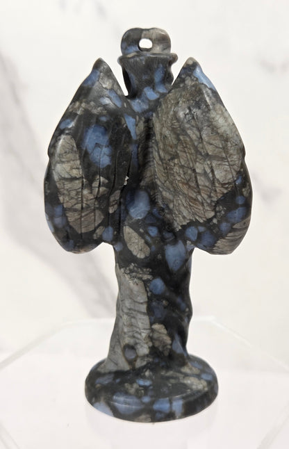A small carved stone angel figurine with wings spread, standing on a clear display base against a light marble-patterned background.