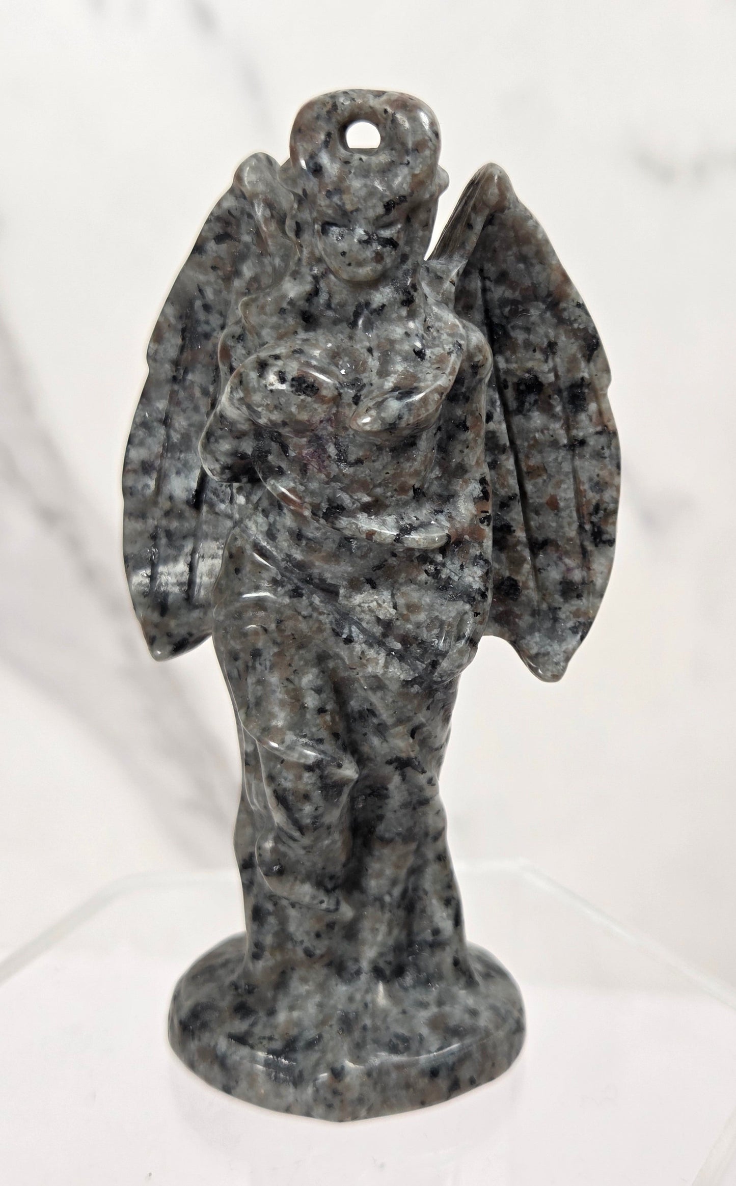 A small carved angel figurine made of speckled gray and black stone, standing on a clear display base against a light marble-patterned background.