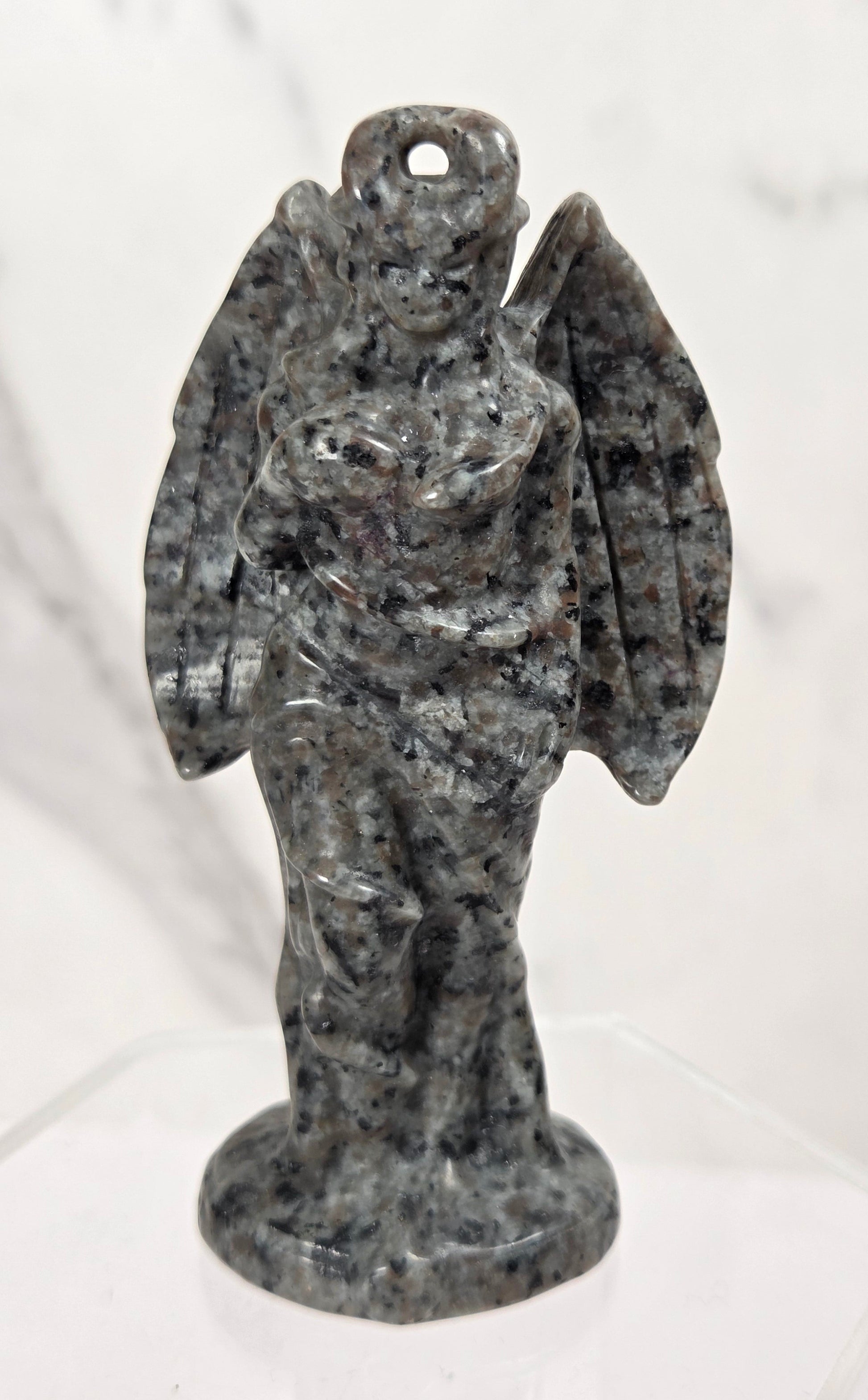 A small carved angel figurine made of speckled gray and black stone, standing on a clear display base against a light marble-patterned background.