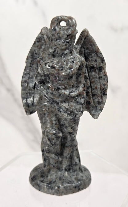 A small carved angel figurine made of speckled gray and black stone, standing on a clear display base against a light marble-patterned background.