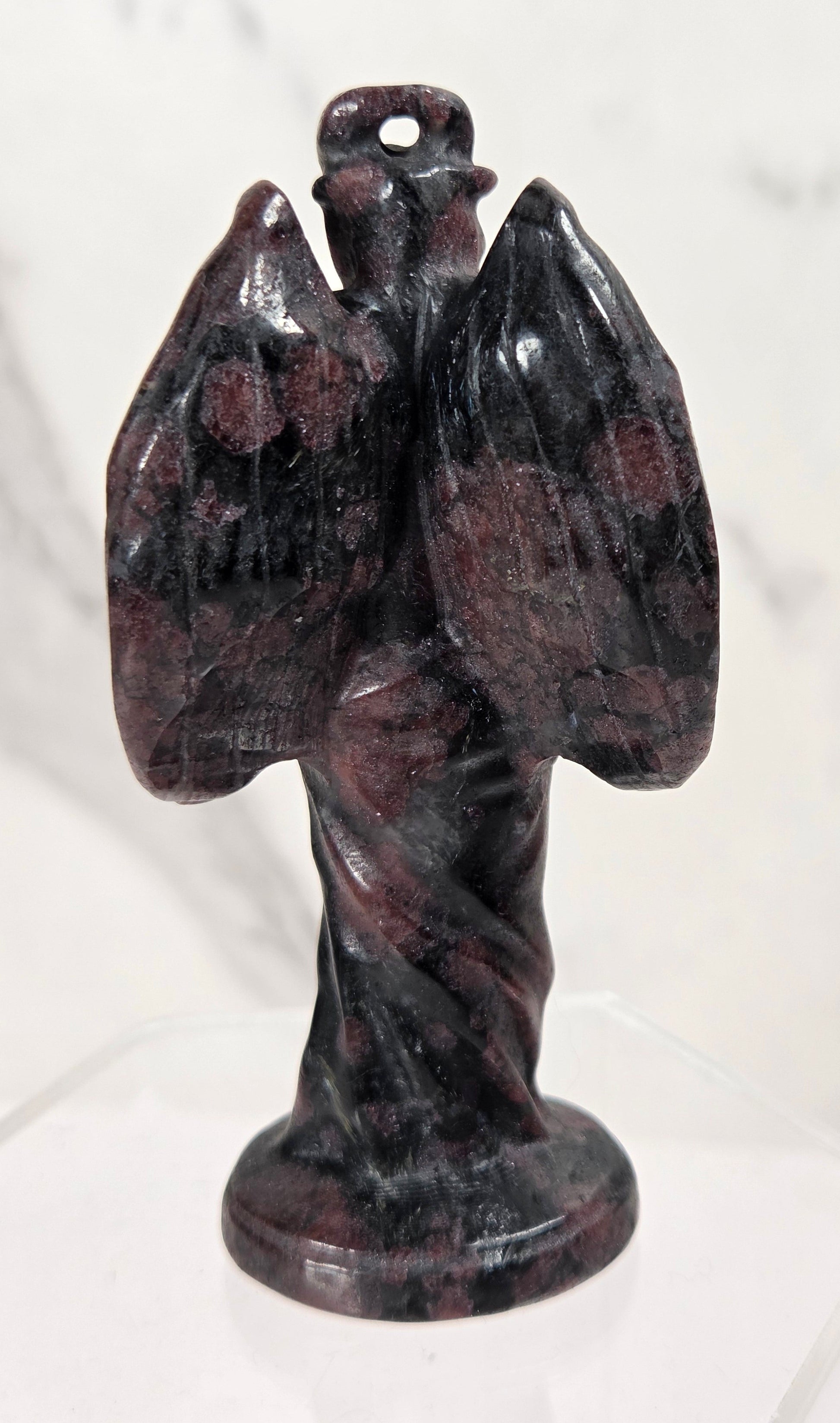 A small carved stone angel figurine with outstretched wings and a loop at the top, displayed on a clear stand against a light marble-patterned background.