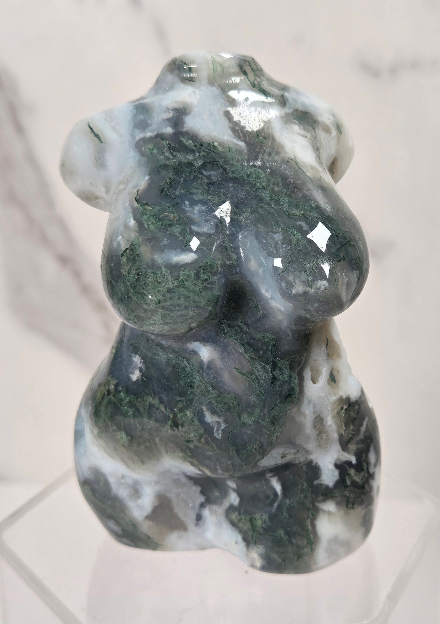 A carved stone figurine of a seated female form, made from polished dark green and white marbled stone, displayed on a clear acrylic stand against a light marble background.