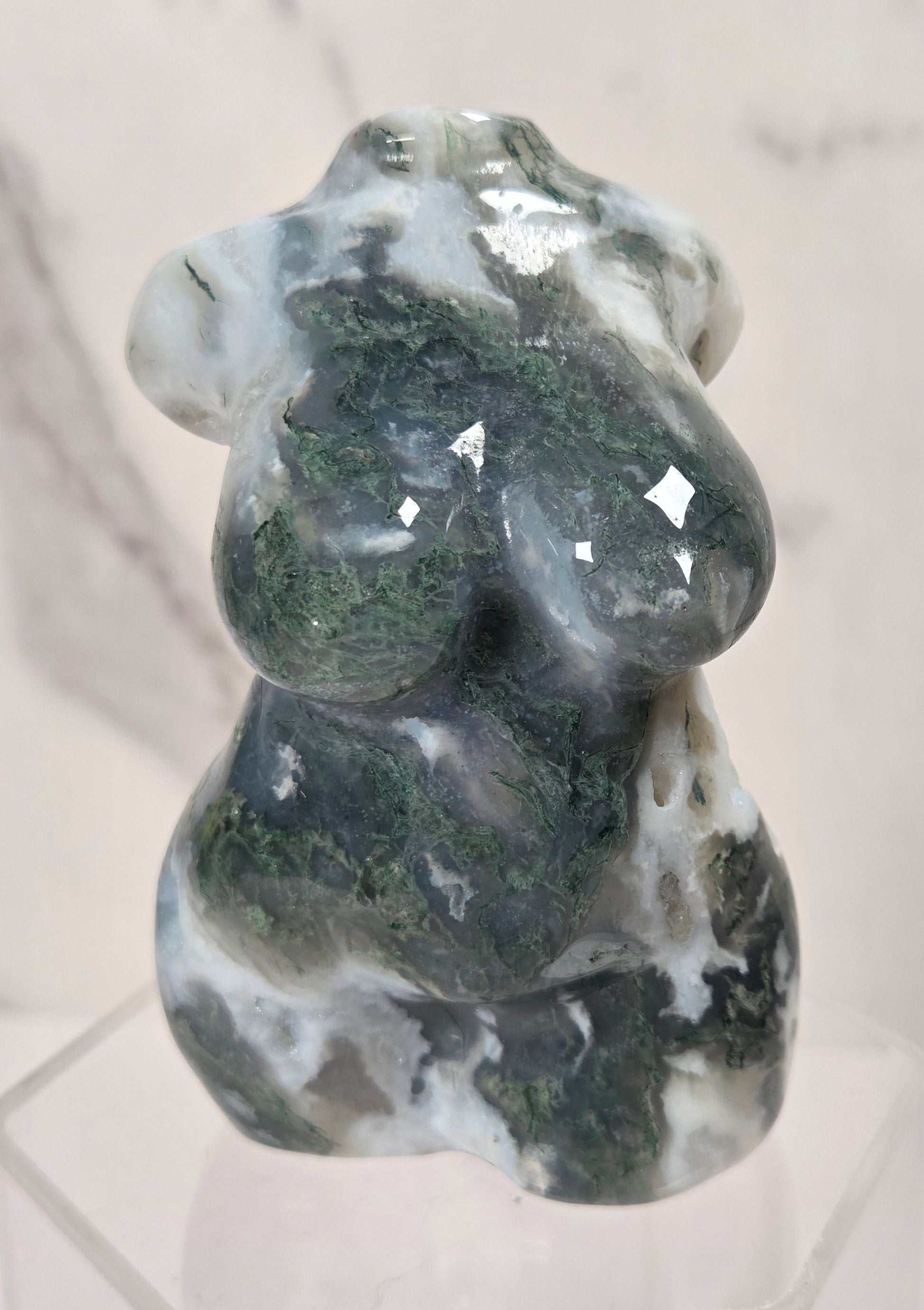 A carved stone figurine of a seated female form, made from polished dark green and white marbled stone, displayed on a clear acrylic stand against a light marble background.