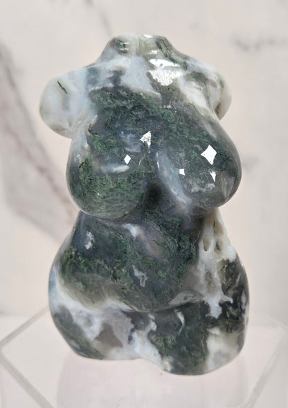 A carved stone figurine of a seated female form, made from polished dark green and white marbled stone, displayed on a clear acrylic stand against a light marble background.