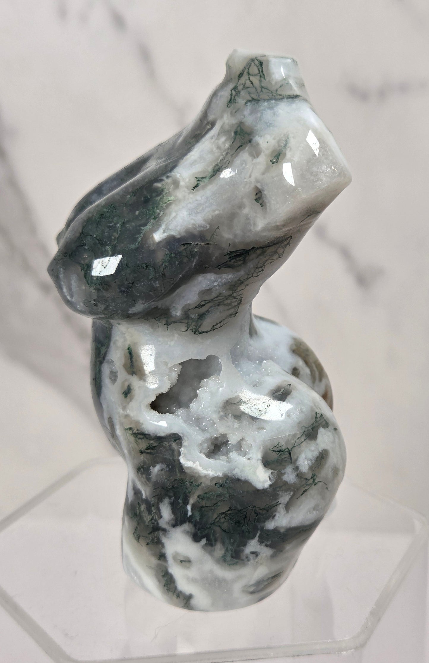 A polished natural stone sculpture with marbled gray, white, and dark green patterns, displayed on a clear acrylic stand against a light marble-patterned background.