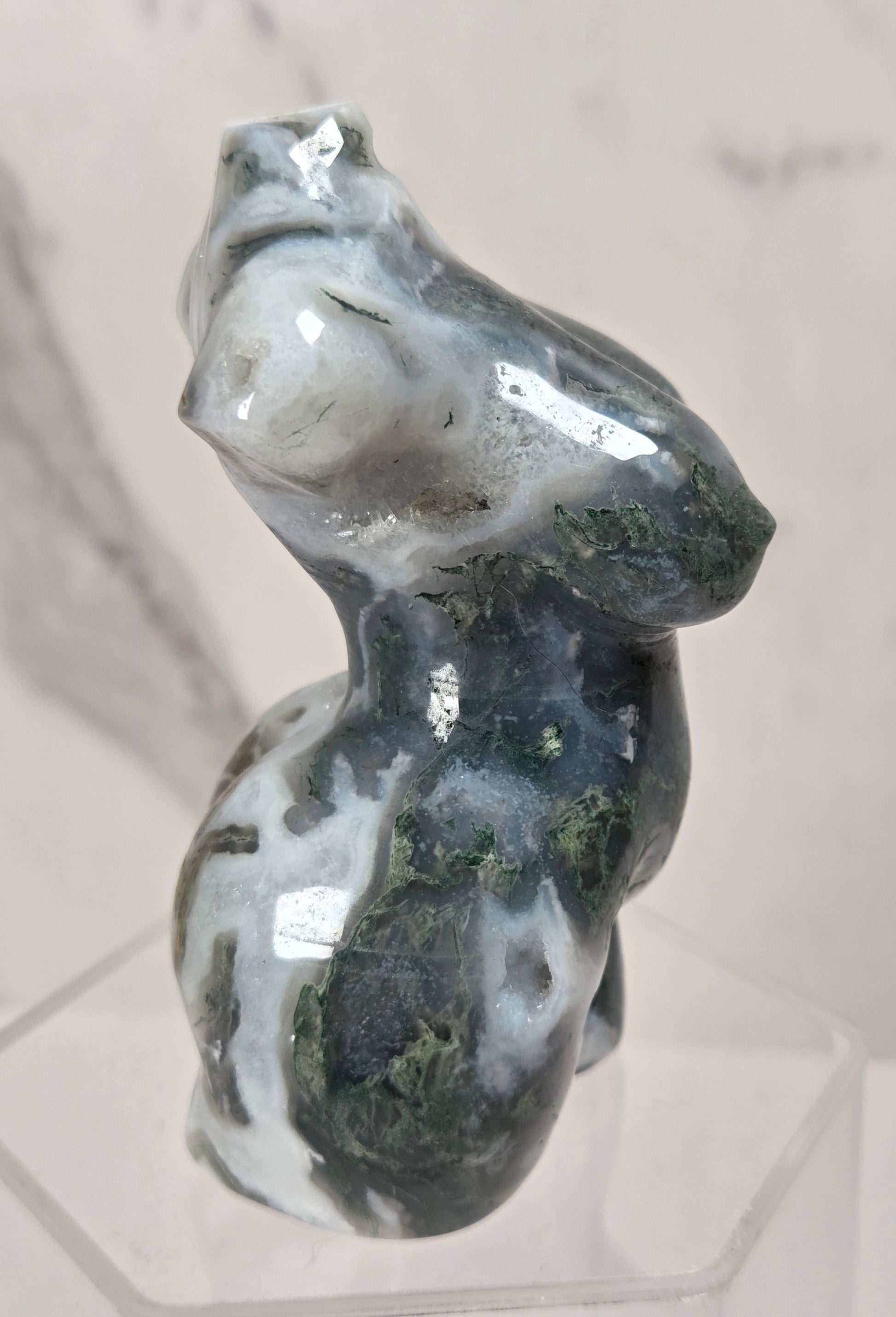 A small sculpted figurine of a seated female form made from polished dark green and white marbled stone, displayed on a clear acrylic stand against a light marble-patterned background.