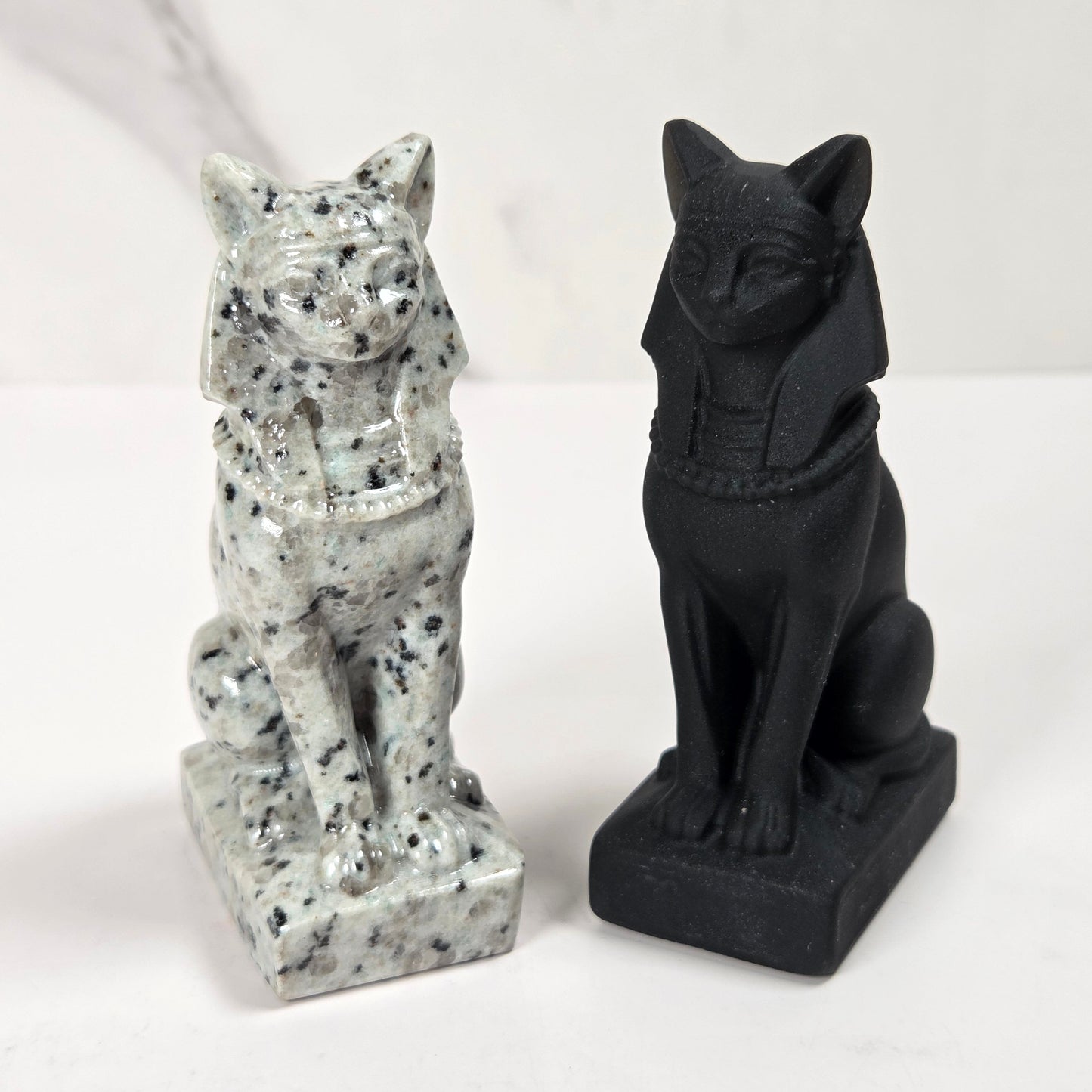 Two small cat figurines shown side by side on a white surface against a light marble-look background. The left figurine is white with black speckling, and the right figurine is solid black.