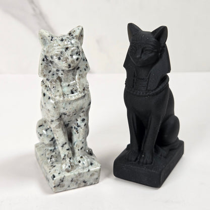 Two small cat figurines shown side by side on a white surface against a light marble-look background. The left figurine is white with black speckling, and the right figurine is solid black.
