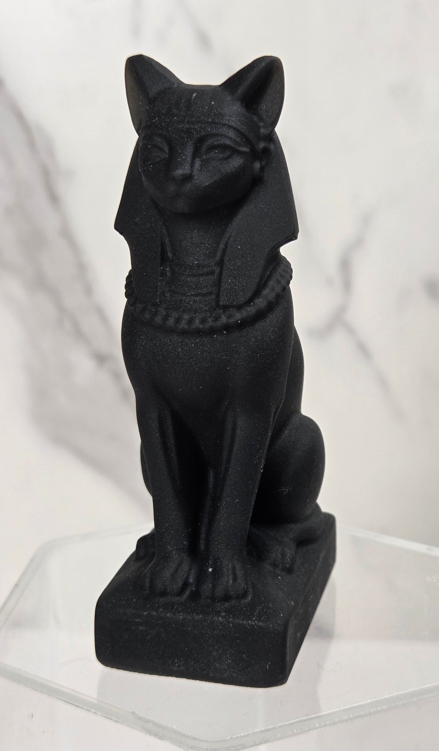 A black Egyptian cat figurine, styled like the Bastet goddess, sitting on a clear acrylic display block against a white marble-patterned background.