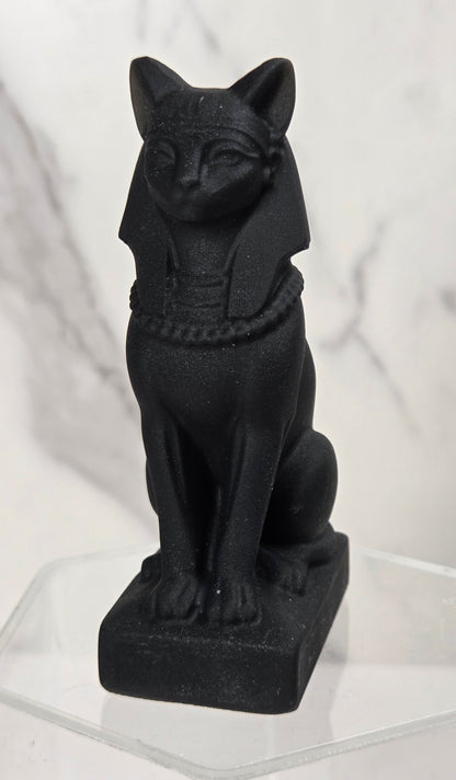 A black Egyptian cat figurine, styled like the Bastet goddess, sitting on a clear acrylic display block against a white marble-patterned background.