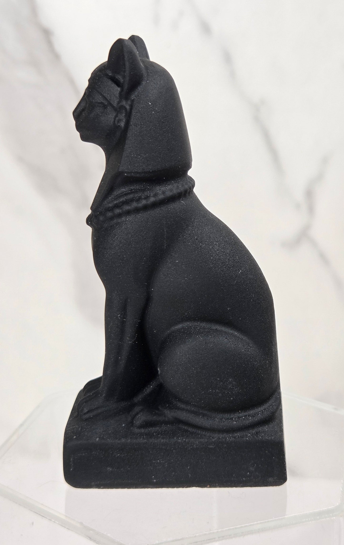 A black matte-finish sphinx statue of an Egyptian cat, displayed on a clear acrylic pedestal against a white marble-patterned background.