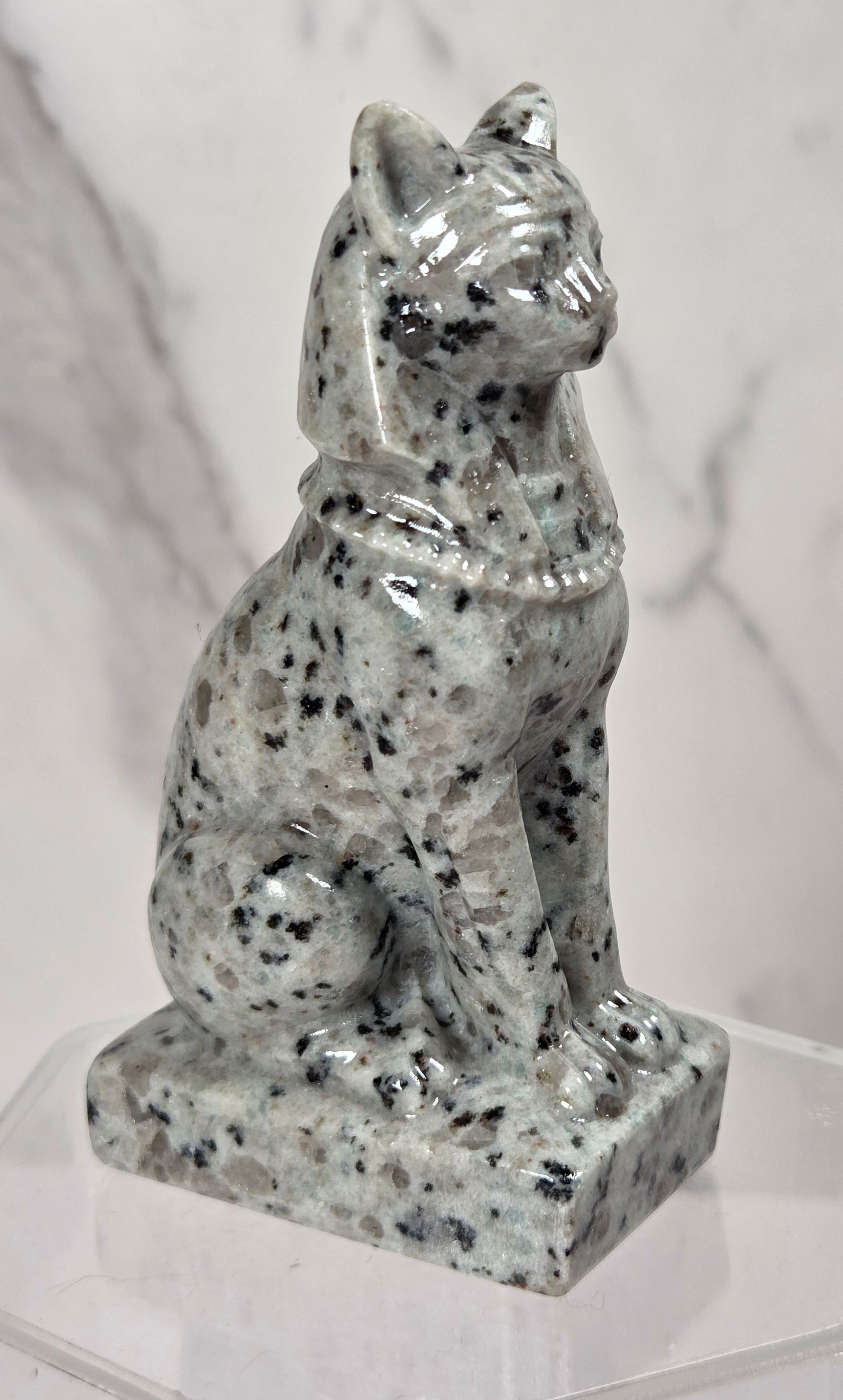 A small seated cat figurine made of speckled gray stone, displayed on a clear acrylic stand against a light marble-patterned background.