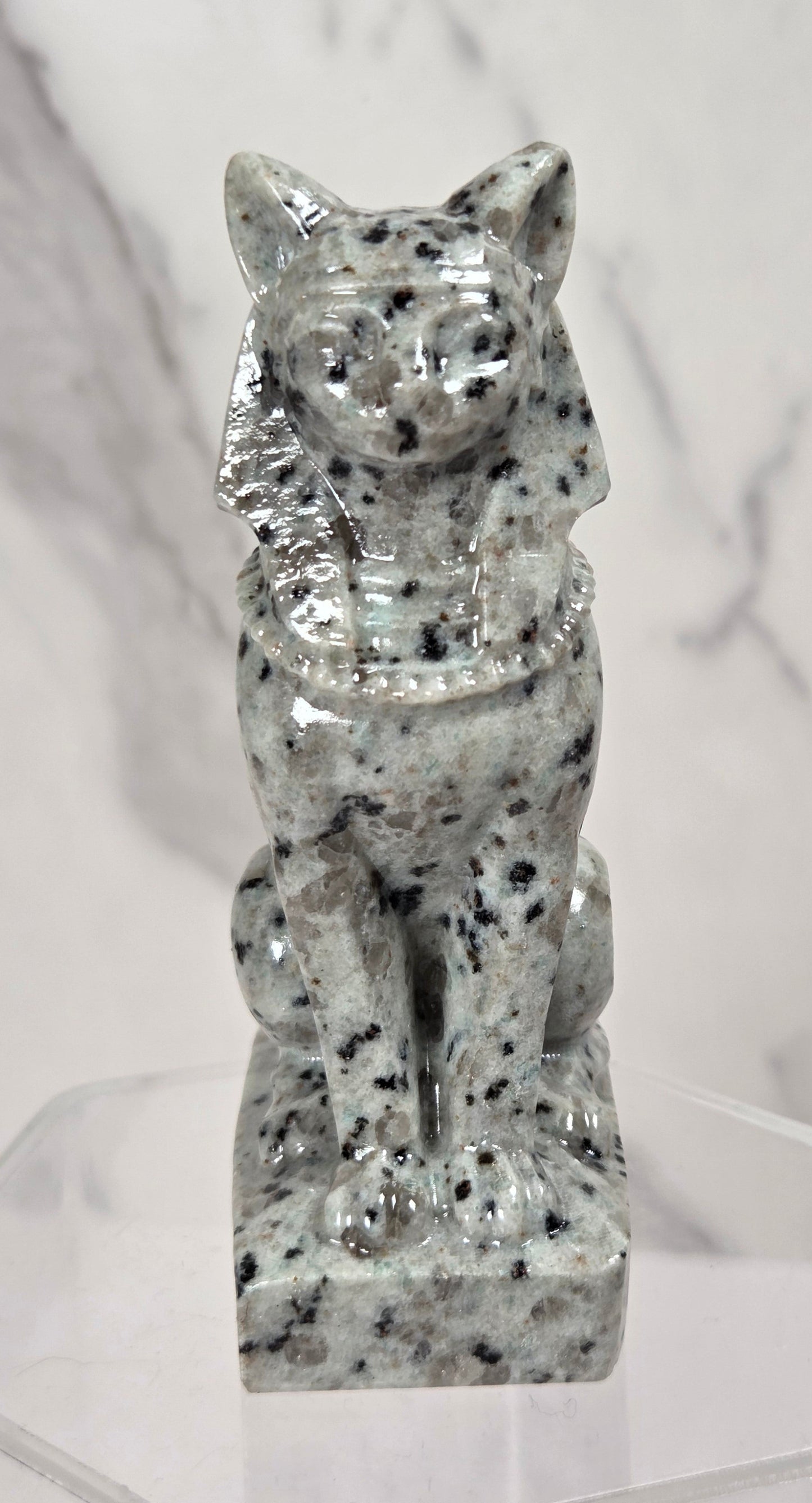 A small glossy stone cat figurine with black and white speckling, displayed on a clear acrylic stand against a light marble-patterned background.