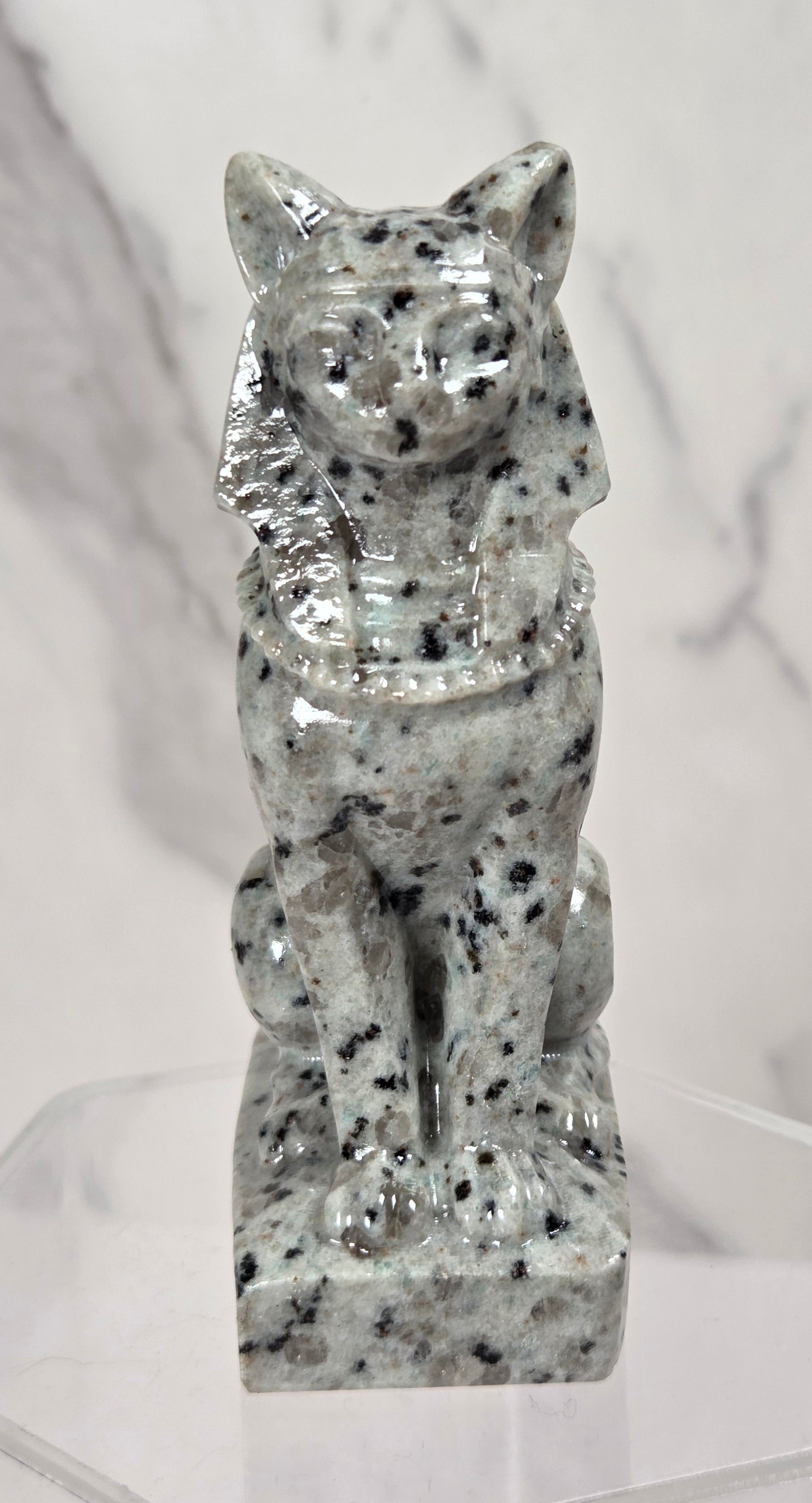 A small glossy stone cat figurine with black and white speckling, displayed on a clear acrylic stand against a light marble-patterned background.