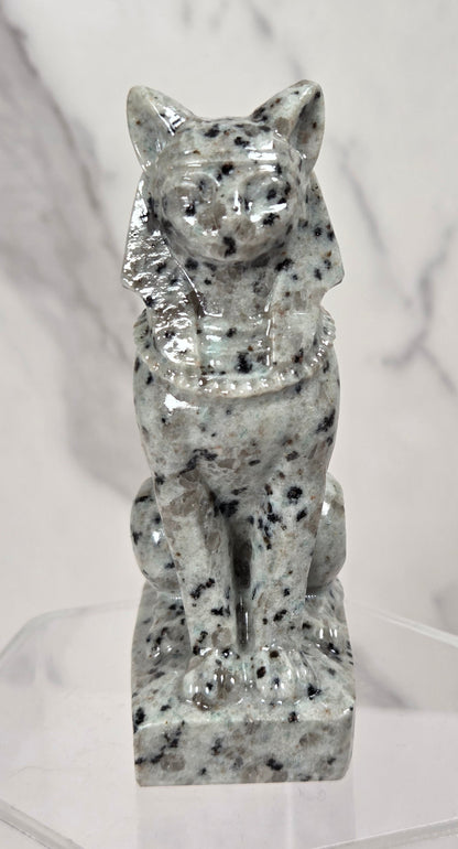A small glossy stone cat figurine with black and white speckling, displayed on a clear acrylic stand against a light marble-patterned background.