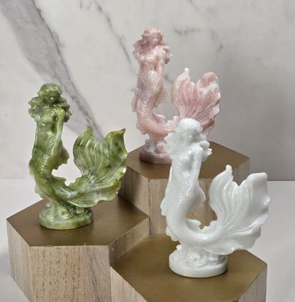 Three small mermaid figurines displayed on hexagonal wooden blocks with metallic gold tops, set against a light marble-patterned background.
