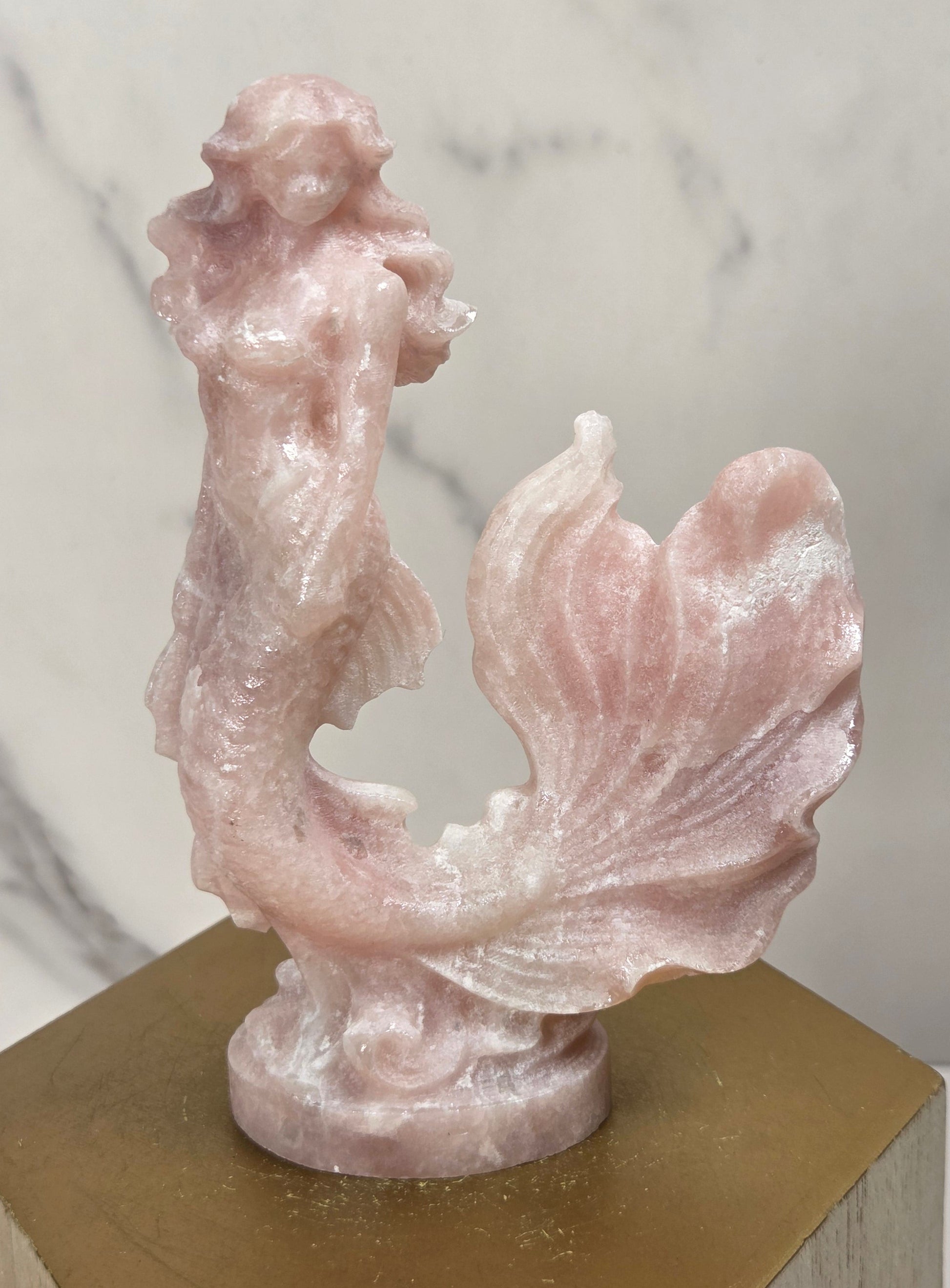 A pink mermaid figurine carved from rose quartz, standing on a gold hexagonal display block against a white marble background.