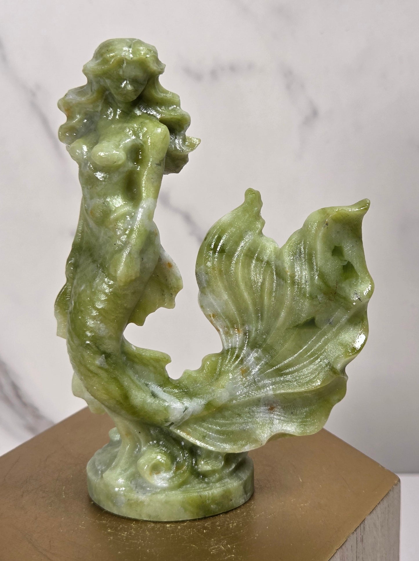 A green jade-like mermaid sculpture standing on a gold-colored hexagonal display block against a white marble-patterned background.