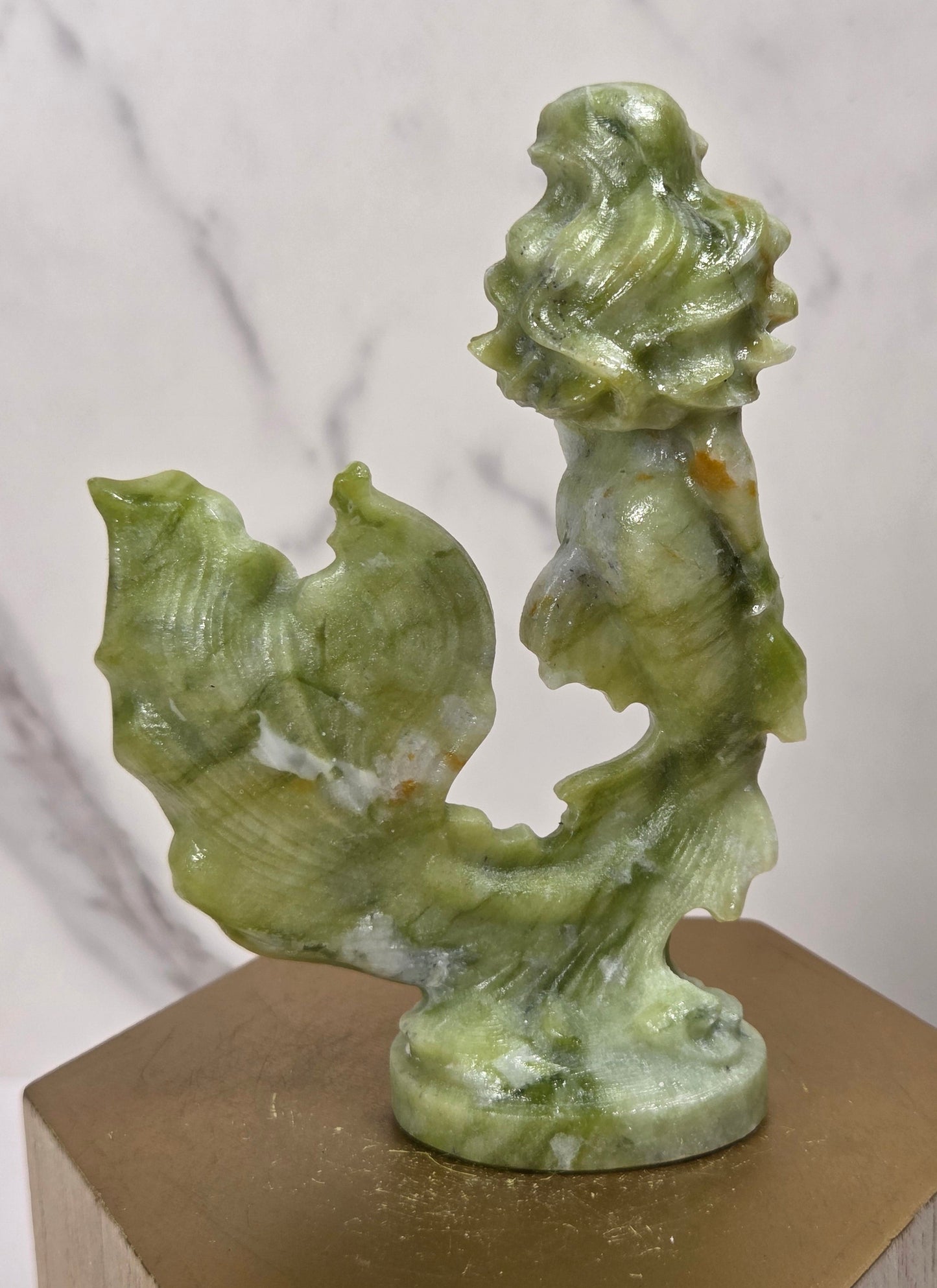 A small green stone sculpture of a mermaid with flowing hair and a tail, displayed on a hexagonal gold-toned pedestal against a light marble-patterned background.