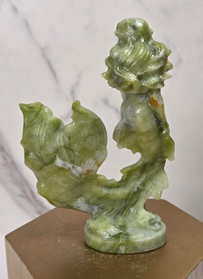 A small green stone sculpture of a mermaid with flowing hair and a tail, displayed on a hexagonal gold-toned pedestal against a light marble-patterned background.