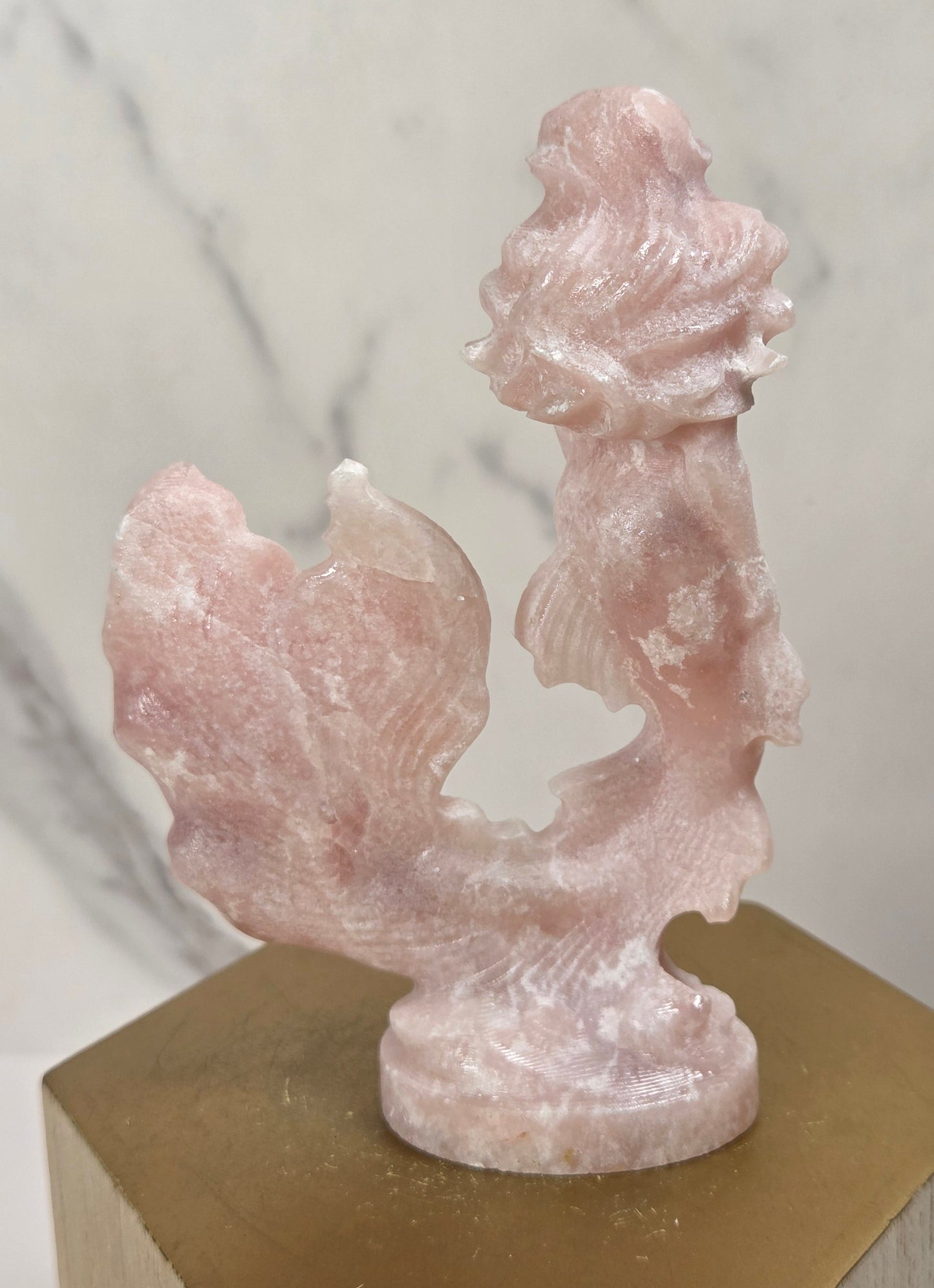 A small carved pink stone sculpture displayed on a gold hexagonal pedestal against a light marble-patterned background.