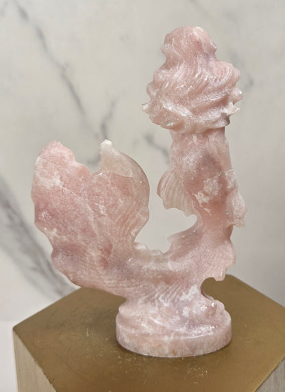 A small carved pink stone sculpture displayed on a gold hexagonal pedestal against a light marble-patterned background.