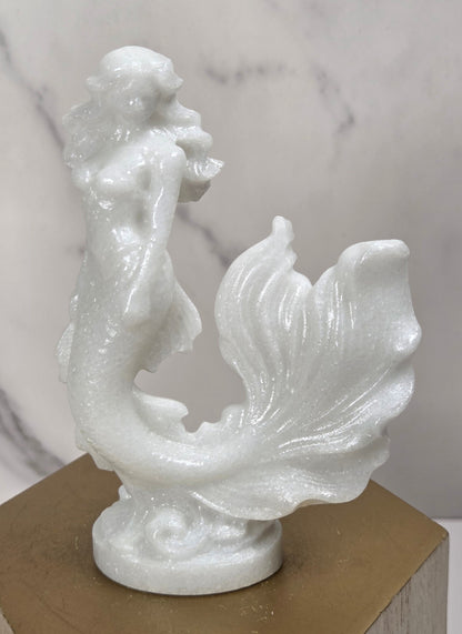 A small white mermaid figurine sculpture displayed on a gold-colored hexagonal pedestal against a light gray marble-patterned background.