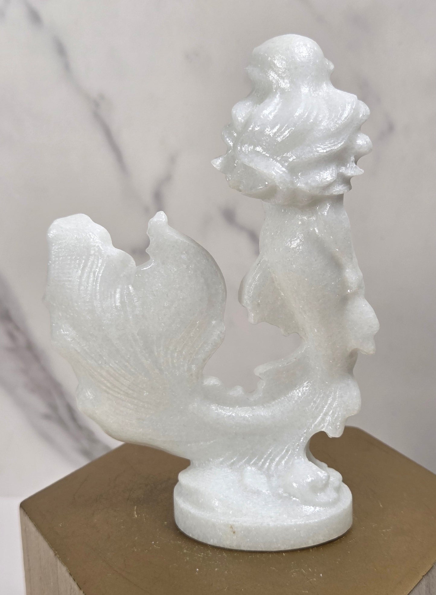 A small white carved figurine of a mermaid with flowing hair and a fish-like tail, displayed on a gold hexagonal pedestal against a light marble-patterned background.