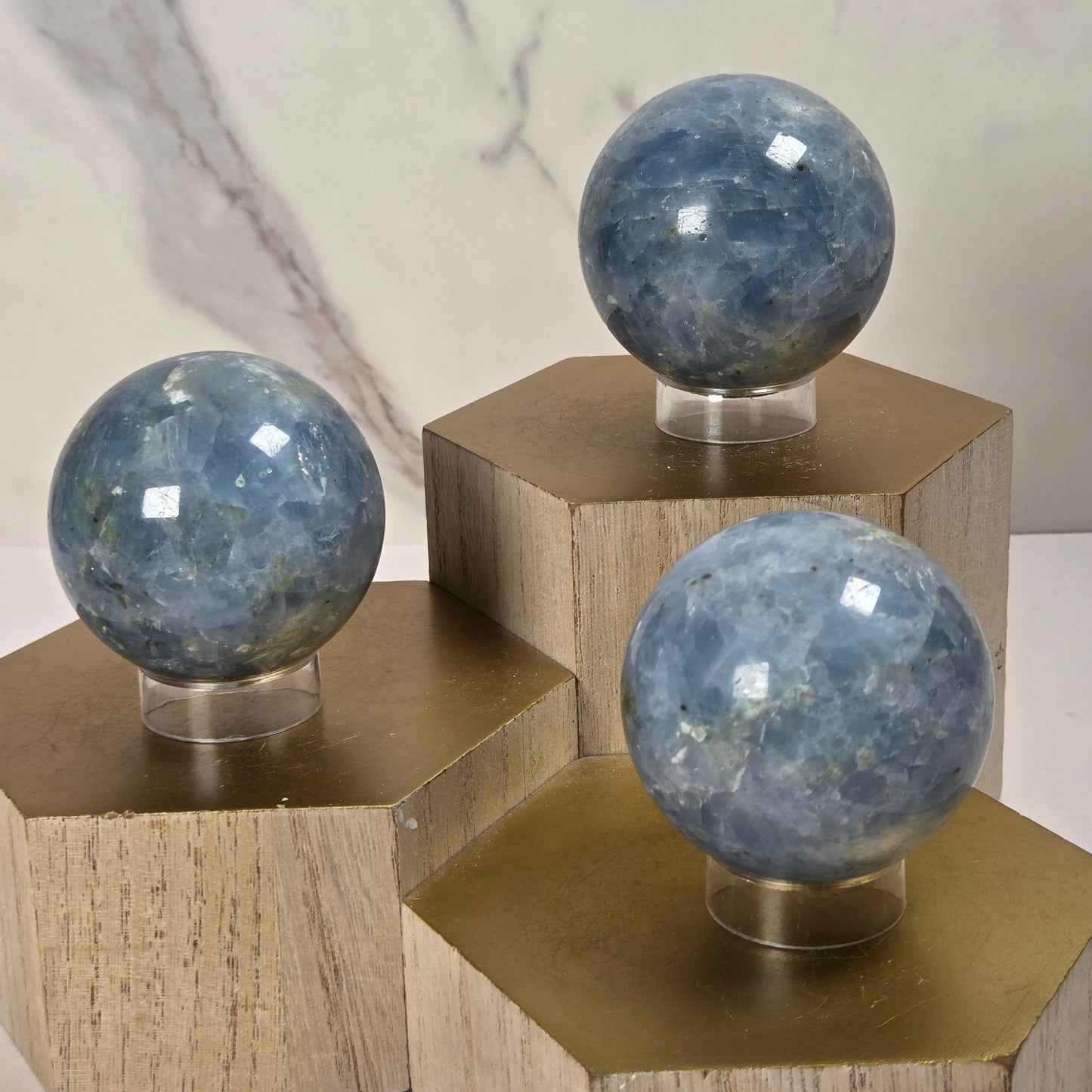 Three polished blue stone spheres displayed on clear stands atop hexagonal wooden pedestals against a light marble-look background.