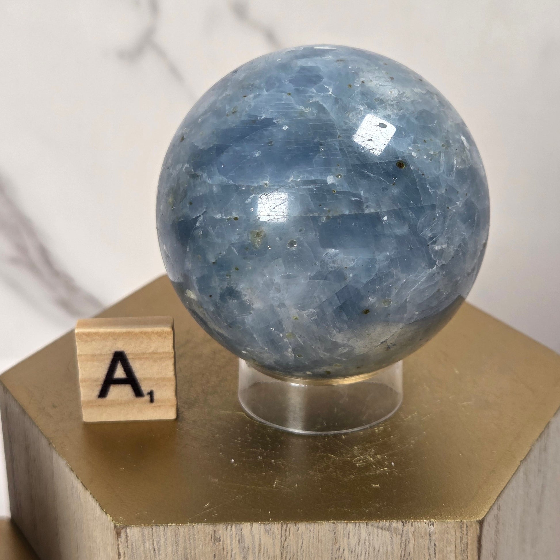 A polished blue-gray stone sphere with natural inclusions, displayed on a clear stand atop a gold hexagonal pedestal, with a small wooden letter block showing 'A.' in front, against a light marble-patterned background.
