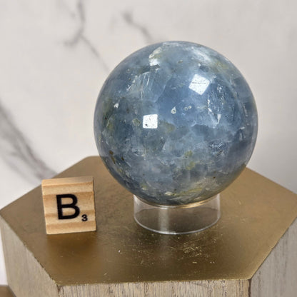 A polished blue fluorite sphere displayed on a clear stand atop a gold hexagonal pedestal, with a small wooden Scrabble tile showing the letter 'B' beside it against a light marble-patterned background.