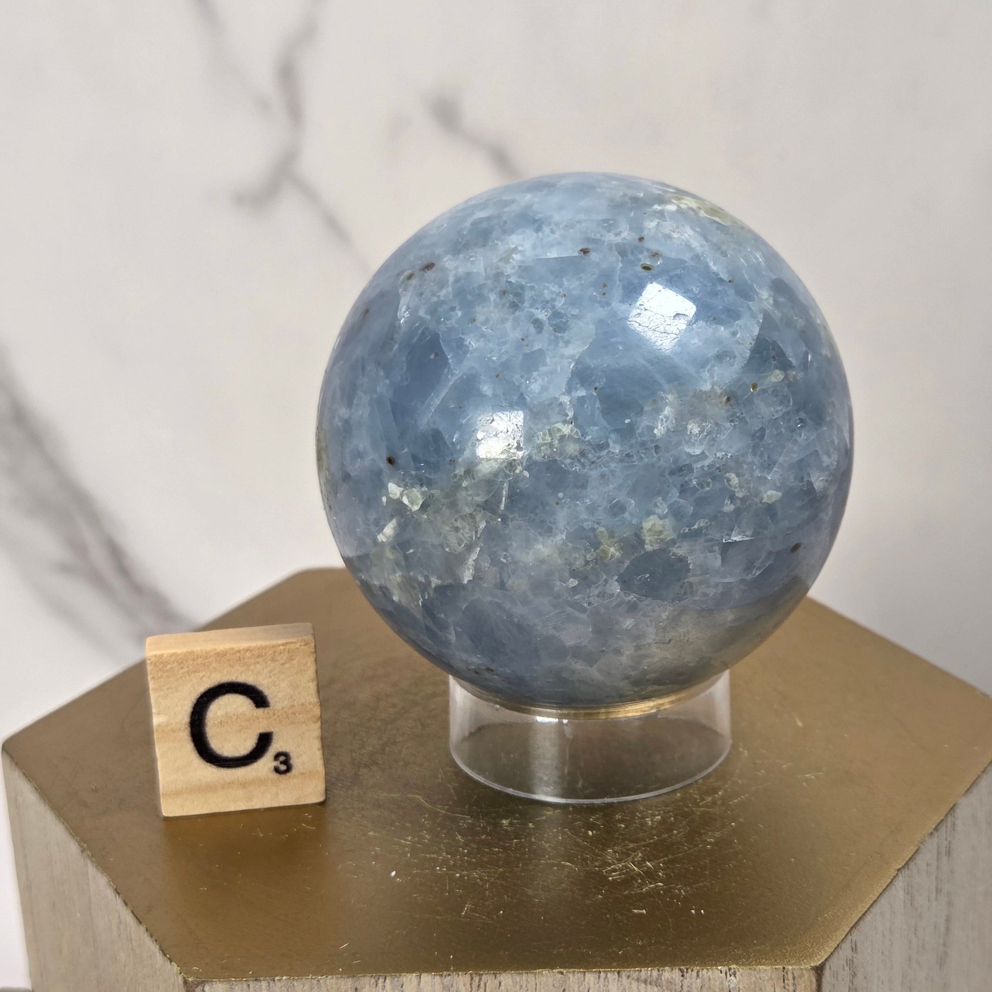 A polished blue stone sphere displayed on a clear stand atop a gold hexagonal pedestal, with a small wooden Scrabble tile showing 'C3' beside it against a light marble-patterned background.
