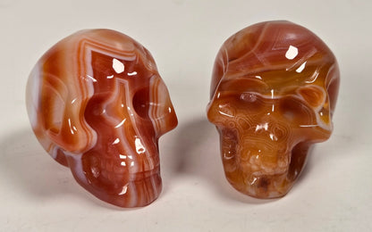 Two polished carved skull sculptures made from banded agate stone, displayed on a light neutral background.