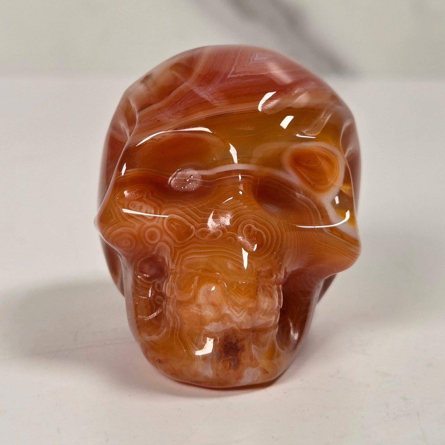 A polished carved skull made of marbled agate or onyx stone in warm amber, orange, and brown tones, photographed on a light neutral surface with a softly blurred marble-like background.