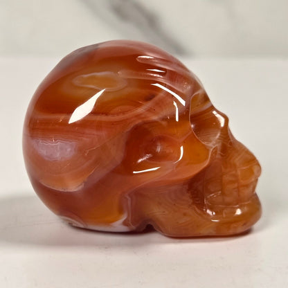 A polished carved skull figurine made of smooth agate or marble, showing warm reddish-brown and amber tones, photographed on a light surface with a soft shadow.