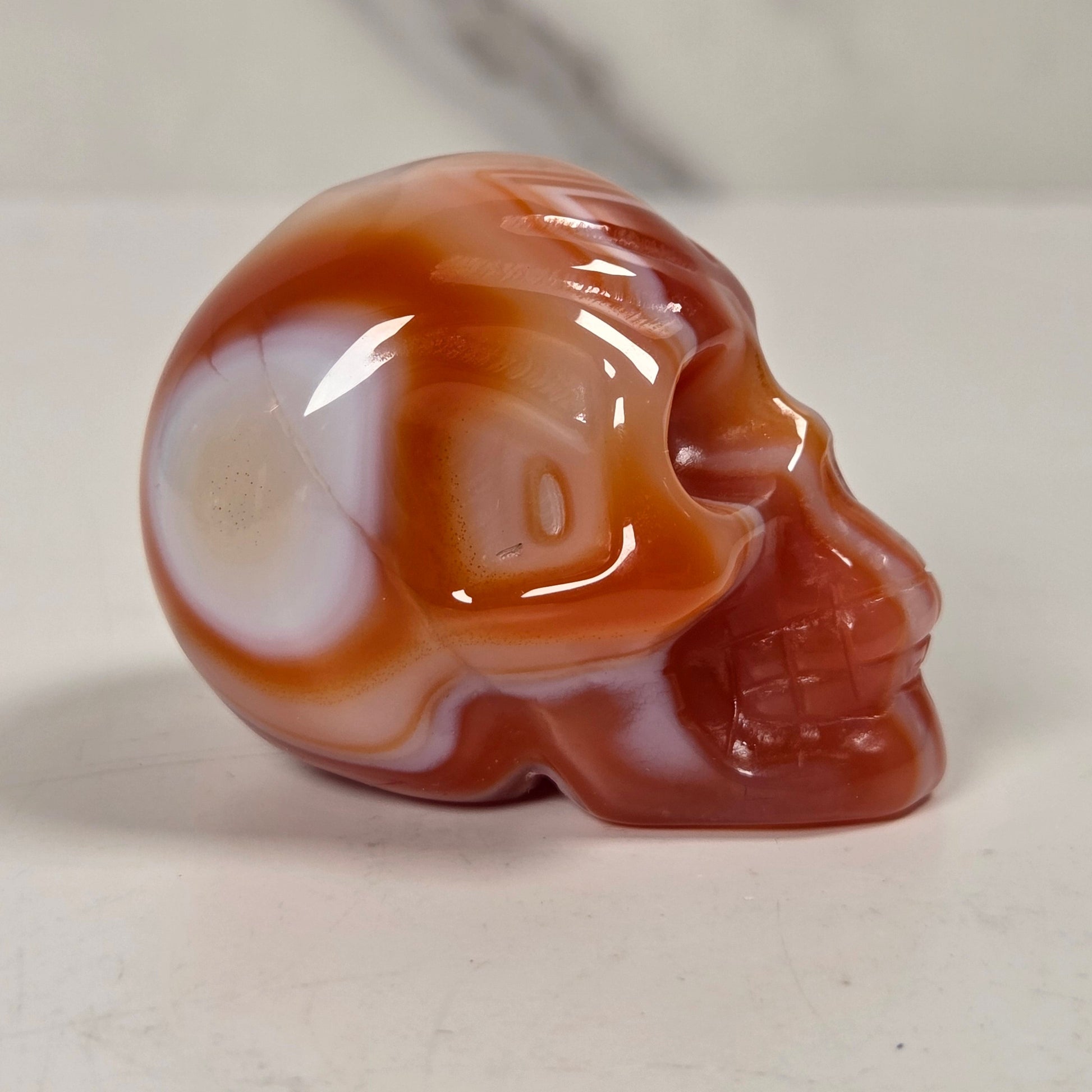 A polished carved skull figurine made of banded agate stone, featuring swirls of reddish-brown, orange, and translucent white, set against a light gray marble-patterned background.