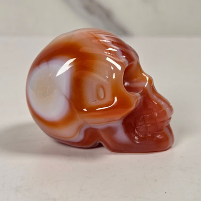 A polished carved skull figurine made of banded agate stone, featuring swirls of reddish-brown, orange, and translucent white, set against a light gray marble-patterned background.