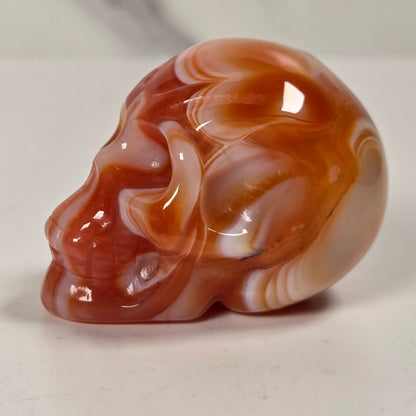 A polished carved skull figurine made of marbled agate stone, featuring swirls of reddish-brown, orange, and cream, set against a light off-white background.