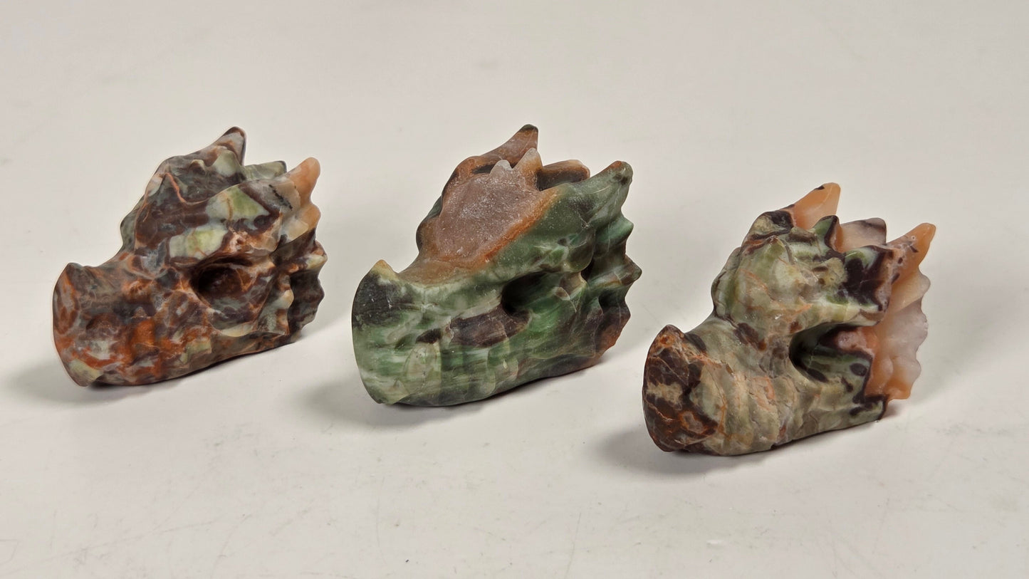 Three small carved stone dragon heads in earthy marbled tones of green, brown, beige, and orange, arranged on a plain light background.