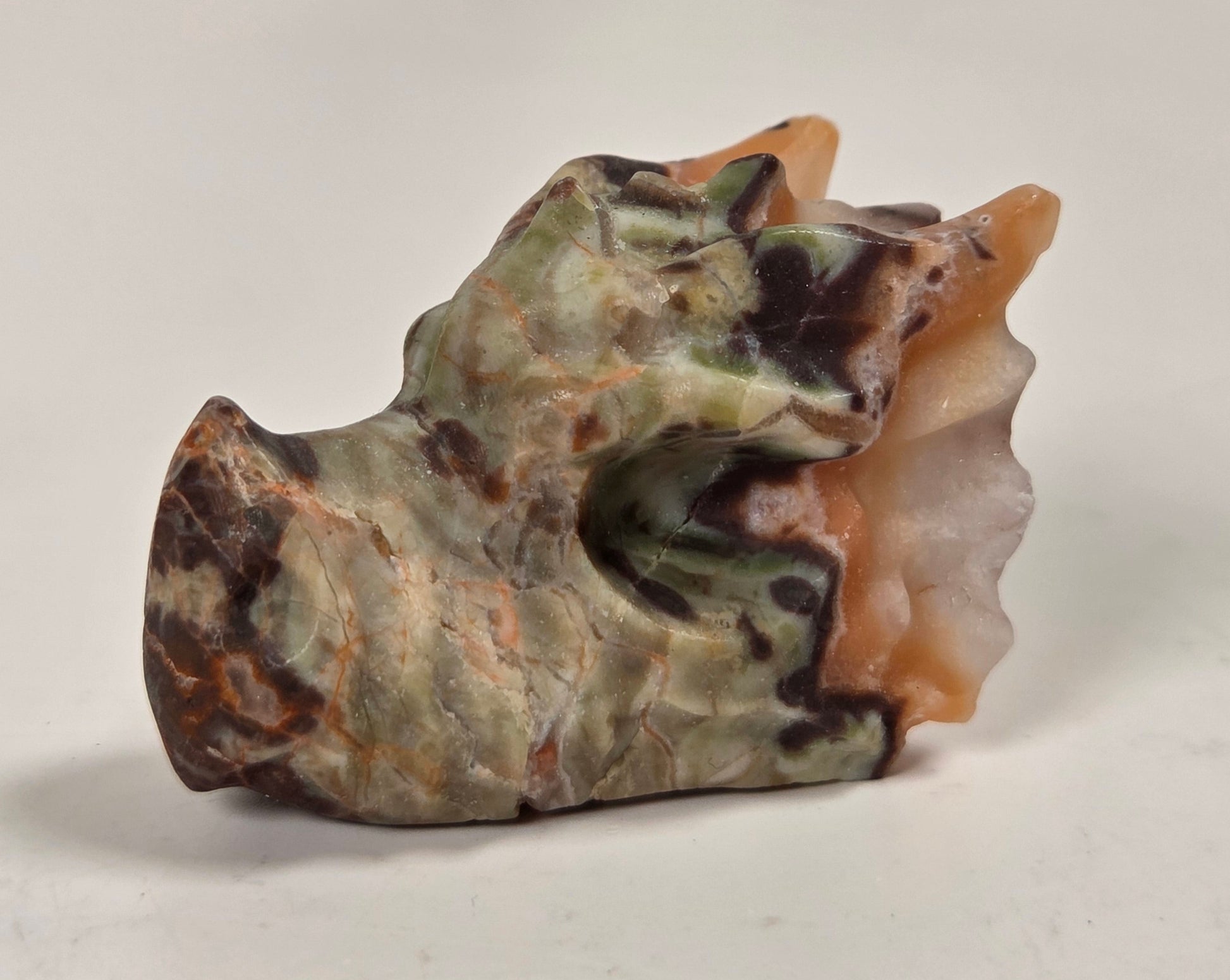 A carved stone sculpture resembling a stylized animal head or horn-like form, made from marbled stone with green, brown, beige, and peach tones, photographed on a plain light background.