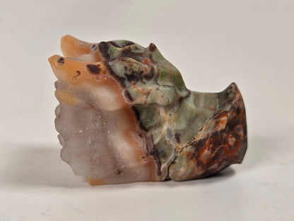 A small carved stone figurine with a dragon-like head and body, featuring natural marbled colors including green, brown, orange, and cream, set against a plain light background.