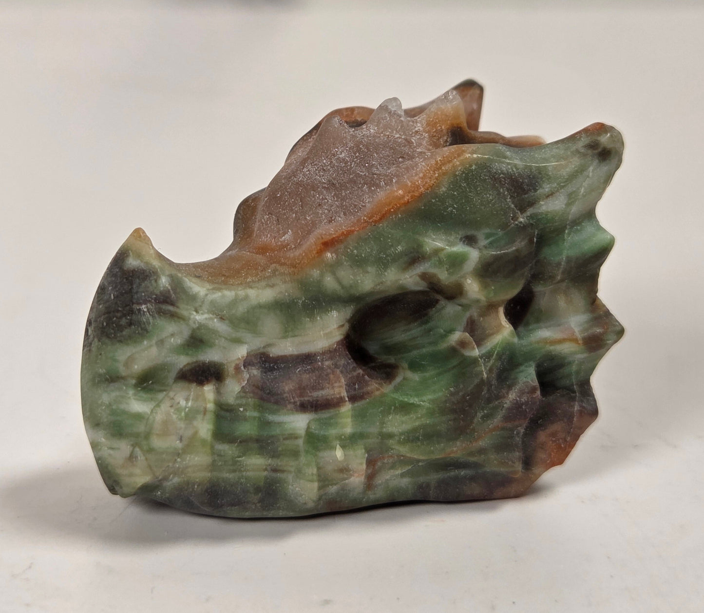 A small carved stone sculpture resembling a stylized animal head or skull, made from green, brown, and gray marbled stone, photographed on a light surface with a blurred neutral background.