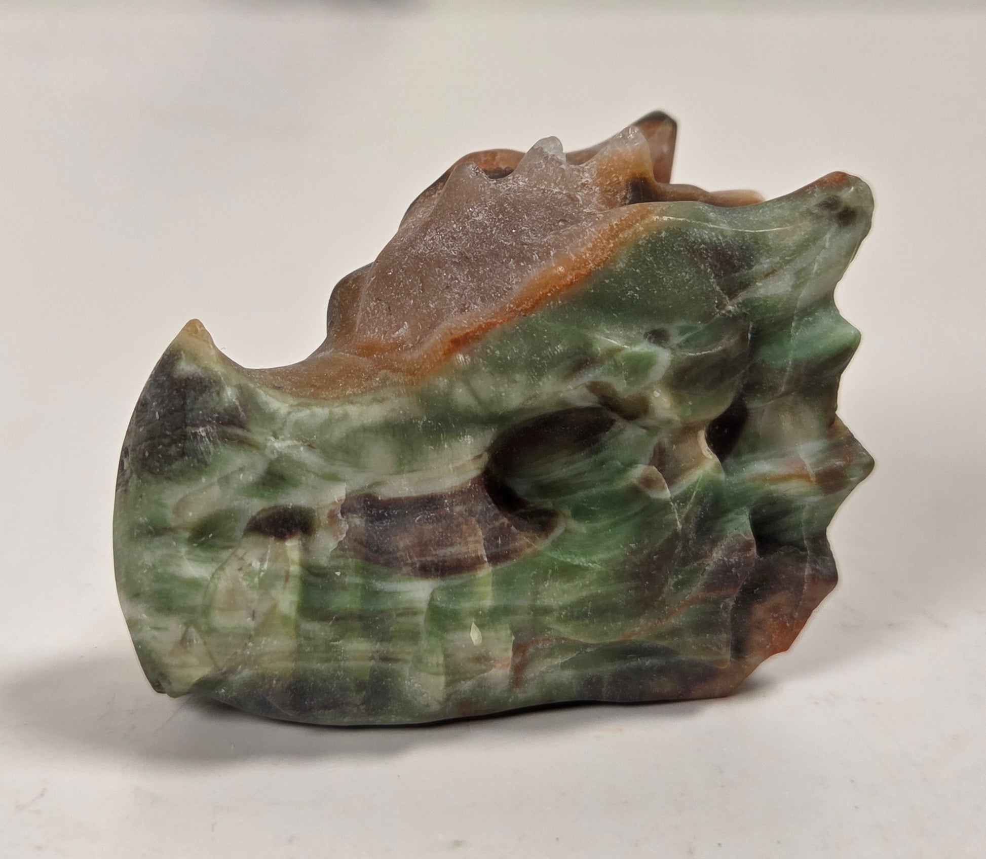 A small carved stone sculpture resembling a stylized animal head or skull, made from green, brown, and gray marbled stone, photographed on a light surface with a blurred neutral background.