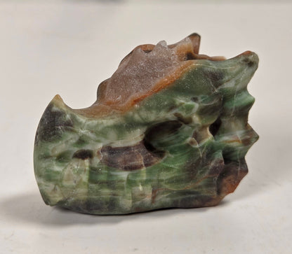 A small carved stone sculpture resembling a stylized animal head or skull, made from green, brown, and gray marbled stone, photographed on a light surface with a blurred neutral background.