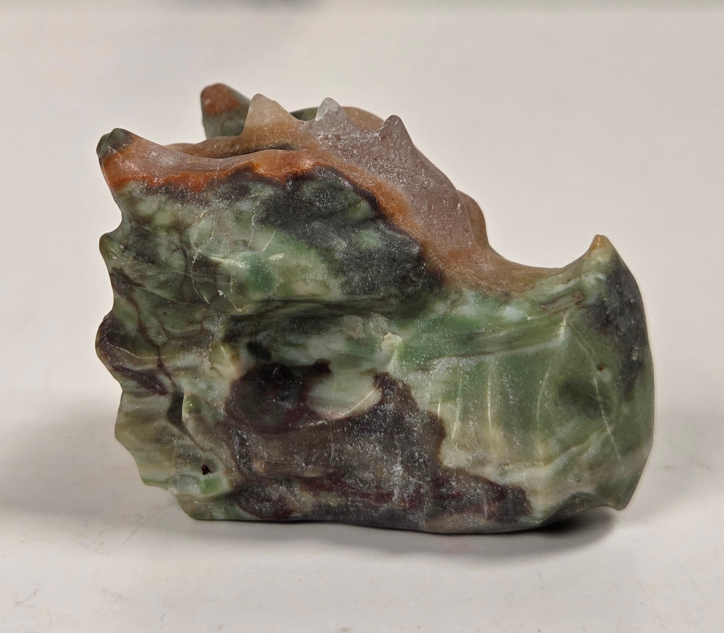 A small carved stone sculpture depicting a stylized landscape or mountain scene, featuring green, brown, and dark gray marbled stone textures, photographed on a light gray surface.